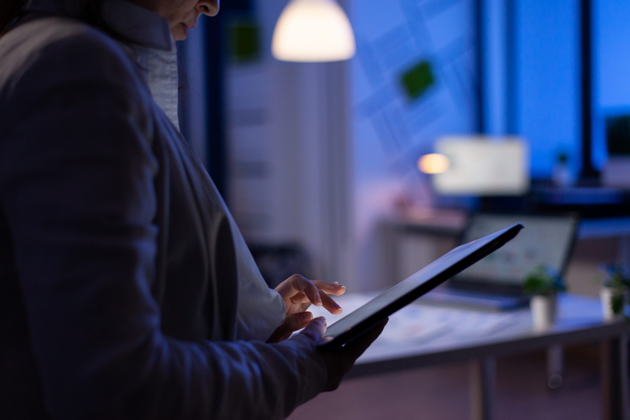Close-up of woman hands typing on tablet checking financial graphs standing in start-up office late at night. Manager using social network, texting and blogging working overtime for job